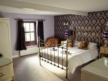 A bedroom with a brass bed white bedding floral pillows two wooden bedside tables with lamps a brown armchair a window with dark curtains and patterned wallpaper at Greety Gate House in Foxfield