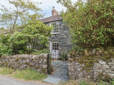 A stone cottage with a white door and window behind a stone wall and iron gate surrounded by bushes at Stair Cottage in Stair nr Keswick Cumbria