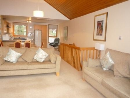 A living room with two beige sofas and a wooden railing with an open kitchen and dining area in the background at 20 Keswick Bridge in Keswick