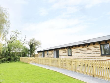 A wooden building with a fenced pathway and green lawn surrounded by trees at Willow Lodge in Cleeve Prior