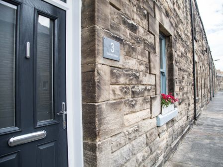 A stone exterior wall with a black door and a window with a flower box at Seashell Cottage in Amble-By-The-Sea
