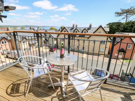 A balcony with two metal chairs and a round table with a bottle and glasses overlooking residential buildings and the sea at 24 Rhodewood House in Saundersfoot