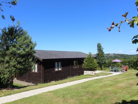 A wooden lodge with a sign reading Willow a paved path green trees lawn and outdoor seating with an umbrella at Willow Lodge Lanivet near Bodmin