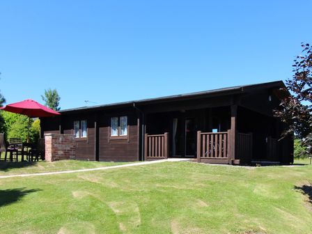 A dark wooden lodge with a porch on a grassy area with an outdoor table and red umbrella at Cherry Lodge in Lanivet near Bodmin