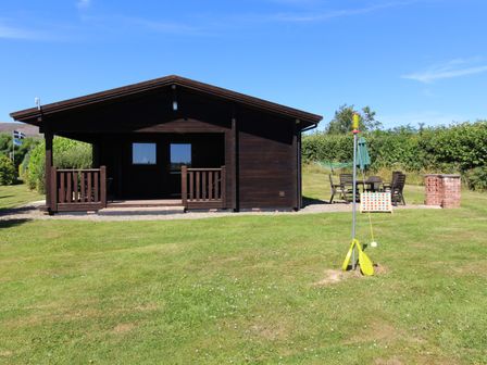 A wooden cabin with a covered porch on a grassy lawn with outdoor furniture and a backyard game at Maple Lodge Lanivet near Bodmin