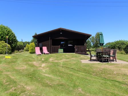 A wooden cabin with two pink chairs and outdoor dining table on a grassy lawn at Cedar Lodge in Lanivet near Bodmin