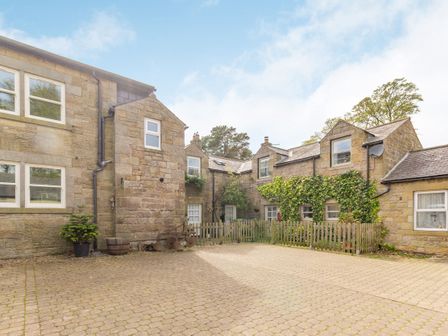 A courtyard with stone buildings and a wooden fence with ivy on one building at Briar Cottage in Edlingham