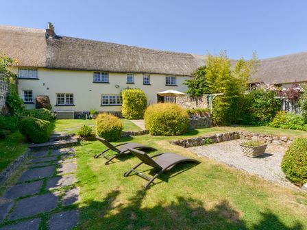A garden with two lounge chairs stone path bushes and a thatched roof house at Michaelmas Cottage in Drewsteignton