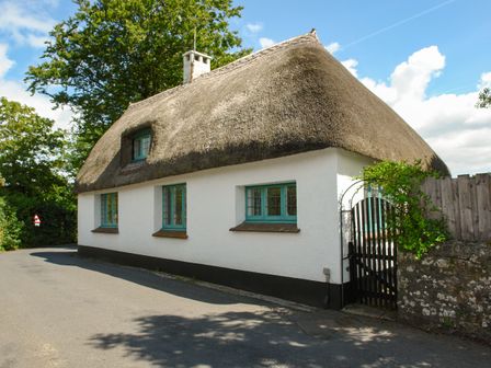 A thatched roof cottage with white walls green windows and a black gate on a street at The Old Forge in Ilsington