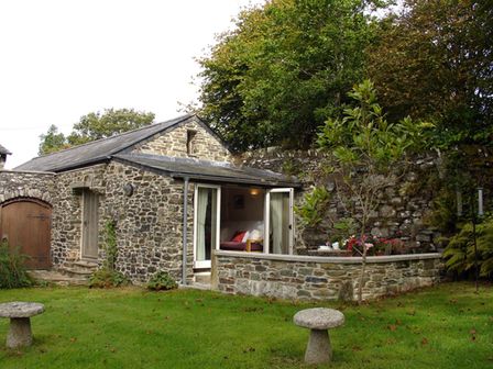 A stone cottage with open doors leading to a garden with green grass and two stone stools at Burham Barn in Dousland near Yelverton