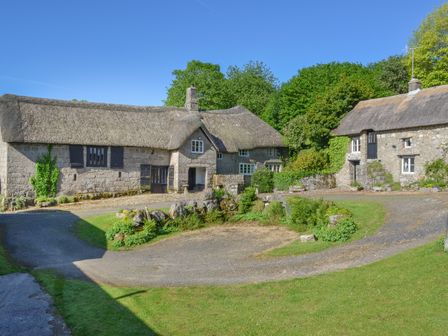 A courtyard with three stone cottages with thatched roofs and surrounding greenery at Hole Farm in Chagford