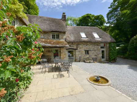 An outdoor patio with metal chairs and table next to a stone cottage with a thatched roof at Bagtor Mill in Ilsington near Haytor Vale