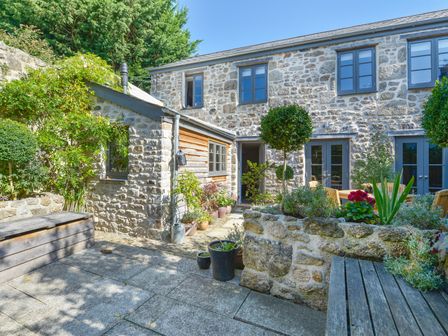 An outdoor stone patio with wooden benches potted plants and a stone planter outside a stone house with multiple windows at Dairy Cottage in Chagford