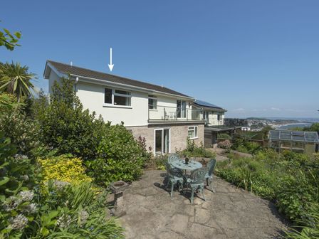 A backyard patio with metal table and chairs surrounded by plants and a two-story house with balcony at Exe View in Dawlish