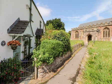 A pathway between a white cottage with plants and a stone church with arched windows at Castle View in Dunster