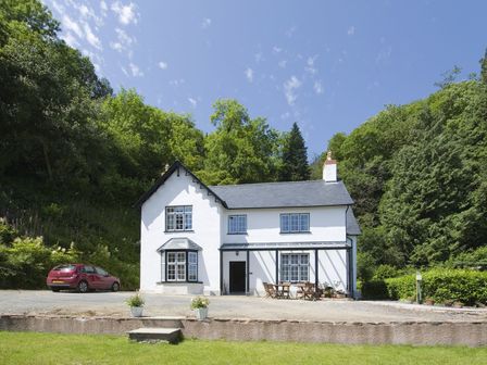 A white two-story house with black window frames and a patio with wooden chairs and table surrounded by trees at The Mount in Dulverton