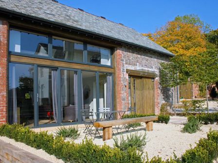 An outdoor patio area with metal chairs and a wooden bench in front of a building with large glass doors at Barley House in Talaton near Ottery St Mary