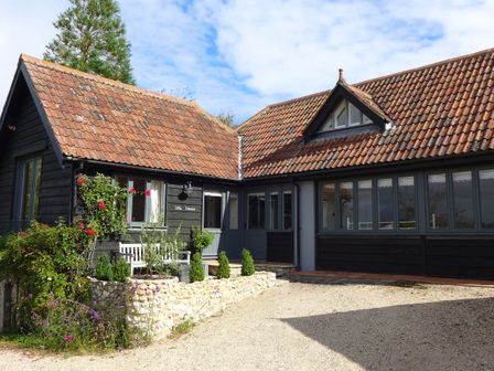 An exterior view of a house with black wooden walls and red tiled roofs with a white bench and plants in front at Little Evanses in Dalwood near Axminster
