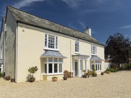 A gravel driveway with potted plants and benches in front of a cream colored house with white framed windows at Abbots Manor in Combe Raleigh near Honiton