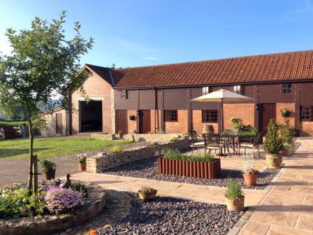 A courtyard with potted plants tables chairs and umbrella outside a barn building at The Linney in Colyton