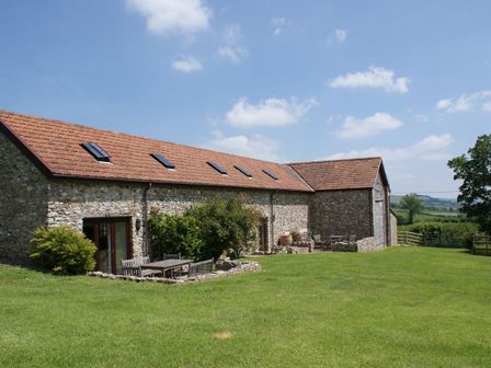 A stone barn with multiple skylights and outdoor wooden tables and chairs on a grassy lawn at Battens in Colyton