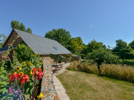 A stone house with a slate roof and garden furniture on a stone patio surrounded by flowers and trees at The Old Parlour in Sampford Courtenay