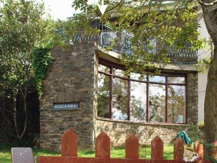 A stone building with large curved windows and a sign reading Galanda behind a wooden fence with trees around at Galanda Treetops Newton Ferrers