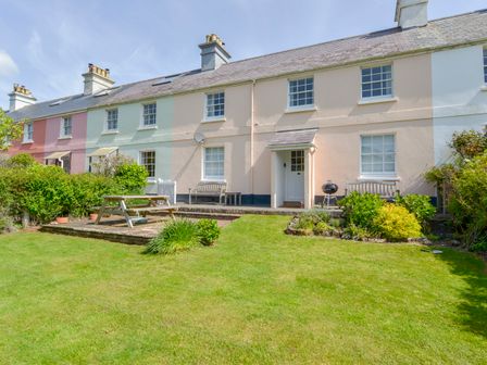 A row of pastel-colored terraced houses with a lawn and picnic bench in the foreground at 4 Coastguards Cottage in Hope Cove