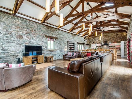 A large open plan living and dining area with stone walls wooden beams leather sofas a dining table and a kitchen in the background at Mill Barn in Woolston near Salcombe