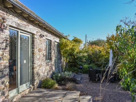 A stone building with glass doors and windows next to a garden area with outdoor furniture and plants at Bradbridge Barn in Newton Ferrers