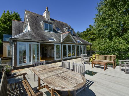 A house with a slate roof and large glass doors opening to a wooden deck with outdoor furniture at Dove Cottage in Dittisham