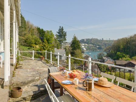 An outdoor terrace with a wooden table and white chairs overlooking a hillside and water at Woodlands in Dartmouth