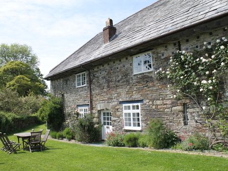 An outdoor garden area with wooden table and chairs beside a stone house with windows and flowering plants at Harry in Blisland
