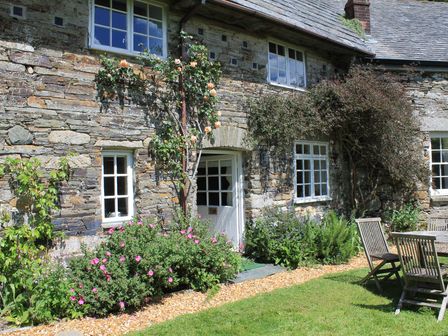 An exterior view of a stone house with white windows a wooden door flowering plants and outdoor wooden furniture on a lawn at Tom in Blisland