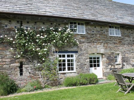 A stone cottage exterior with white windows and door and a garden with climbing plants and wooden table and chairs at Dick Cottage in Blisland