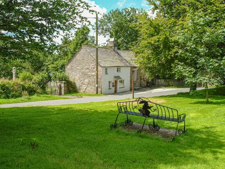 A green lawn with a metal bench and a stone house across a road surrounded by trees at Churchgate Cottage in Blisland