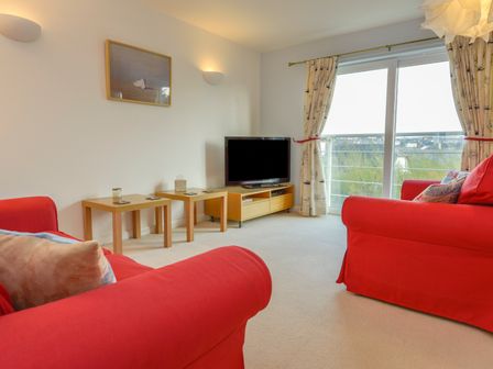 A living room with red sofas a television on a wooden stand two small wooden tables and sliding glass doors with curtains at The Loft in Bude