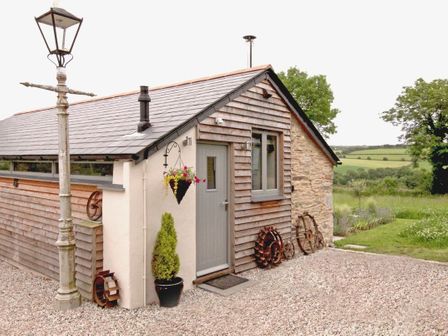 A small building with wooden and stone walls a grey door a hanging flower pot a potted plant and old metal wheels outside Mugwell Barn Luckett near Callington