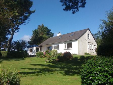 A white house with a dark roof and skylights surrounded by trees and a lawn with a bench at Blue Seas in St Mawes