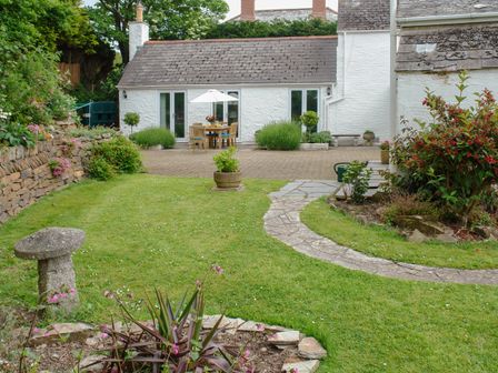 A garden with a stone path, potted plants, a stone bench, and a patio with outdoor dining furniture and an umbrella at Little Barn Cottage in Portloe