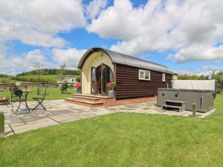 A small wooden cabin with arched roof and open double doors on a paved patio with a hot tub and metal outdoor furniture