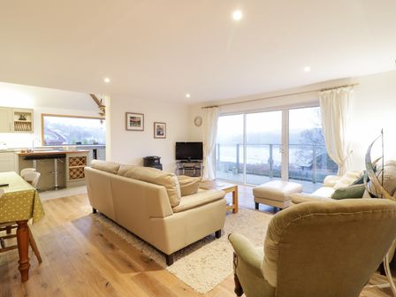 A living room with beige sofas a green armchair wooden floor and sliding glass doors opening to a balcony at Treetops in Golant near Fowey