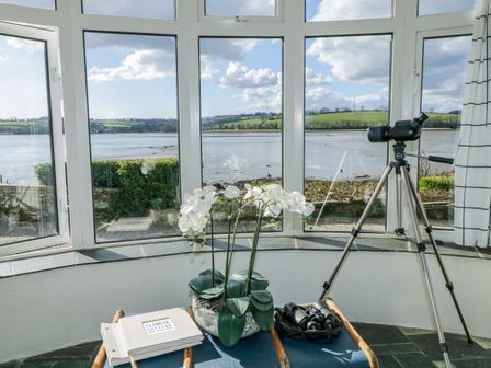 A bay window with a telescope on a tripod a plant a book and binoculars overlooking a river and green hills at Clamoak Cottage in Clamoak near Bere Alston