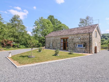 A stone building with a pitched roof surrounded by gravel and a small lawn with young trees and shrubs at Cilgwyn Bach in Llandysul