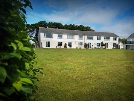 A long white two-story building with several windows and doors overlooking a large grass lawn with trees in the background at Driftwood in Bude