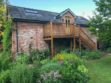 A brick house with a wooden balcony and stairs surrounded by a garden with flowers and plants at The Hayloft in Much Cowarne near Bishop's Frome
