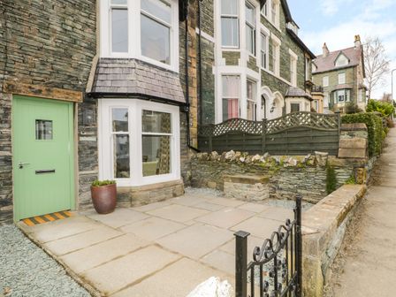 The front exterior of a stone house with a green door a bay window a stone patio and a small fenced garden area at Roselea in Keswick
