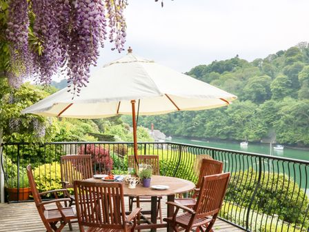 A wooden outdoor dining table with chairs and a large white umbrella on a balcony overlooking a river and green trees at Elphinstone in Newton Ferrers near Plymouth