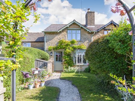 A stone cottage with a gravel path leading to a green door surrounded by plants and garden furniture at Puffitts Cottage in Bourton-on-the-Water