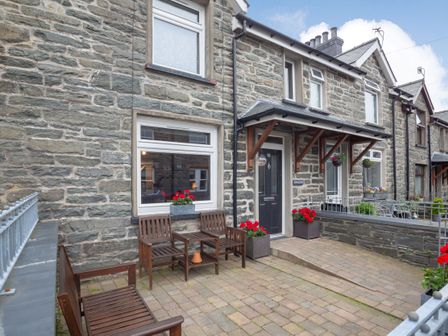 Stone exterior of a terraced house with a black door and windows and a small paved patio with wooden chairs and flower pots at Garreg Ddu in Blaenau Ffestiniog